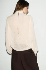 Camisa de cuello redondo y manga larga con puños, confeccionada en tejido semitransparente con textura de pequeños puntos en relieve. Presenta volantes decorativos en el frente y cierre frontal con botones ocultos.