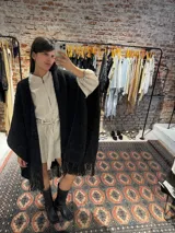 Ruana tejida a mano en lana merino color beige, con textura de punto y detalle de flecos en el ruedo. Presenta sutiles destellos brillantes integrados en el tejido.