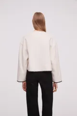 Chaqueta corta de paño color marfil, con cuello redondo, cierre frontal de botones negros y dos bolsillos frontales. Presenta detalles de costuras decorativas en contraste negro en los bordes de los bolsillos, puños y ruedo.