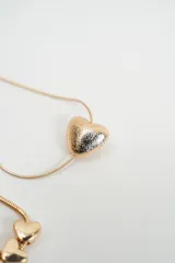 Set de tres pulseras doradas de cadena fina con cierre de mosquetón y cadena de alargue. Incluye una pulsera de cadena trenzada, una pulsera de cadena tipo serpiente con tres dijes de corazones pequeños y una pulsera de cadena tipo serpiente con un dije de corazón grande texturizado.