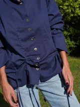 Camisa negra de lino con cuello camisero, abotonada al frente y mangas largas con puños abotonados. Tiene un sistema de ajuste con cordones en la parte delantera que permite usarla larga o fruncida.