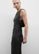 Vestido midi de tejido fluido con estampado de lunares blancos sobre fondo negro. Presenta un diseño entallado sin mangas con escote redondo, tirantes anchos y un detalle de fruncido lateral con lazo que crea un efecto drapeado. Incluye cierre de cremallera invisible en la parte posterior.