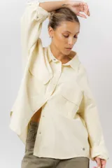 Sobrecamisa de gabardina liviana color crema, con corte oversize, cuello clásico, cierre frontal con botones a presión y dos bolsillos delanteros con solapa.