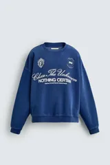 Sudadera de corte boxy fit en color azul con efecto lavado, cuello redondo y manga larga. Presenta estampado frontal con tipografía variada y gráficos estilo universitario. Interior afelpado y acabados en rib.