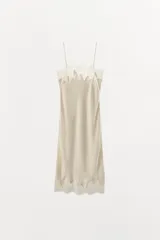 Vestido midi de estilo lencero confeccionado en tejido satinado color beige. Presenta escote recto con tirantes finos y aplicaciones de encaje a tono en el escote y en el ruedo.