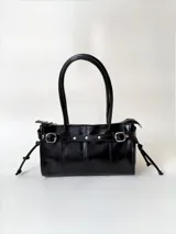 Cartera de cuero negro de estructura rígida, con diseño de tira decorativa con tachas y hebillas metálicas. Cuenta con asas para llevar al hombro, cierre superior y lazos decorativos en los laterales.
