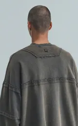 Buzo de cuello redondo, color gris oscuro con efecto desgastado (acid wash). Presenta costuras expuestas y detalles de roturas en los bordes. El calce es oversize y crop, con mangas largas abullonadas y puños y ruedo con terminación deshilachada y ondulada.