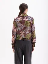 Blusa de manga larga con estampado floral y geométrico en tonos marrones, con cuello clásico, cierre frontal con ganchos metálicos y detalle de volados laterales en la tapeta.
