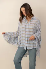 Camisa de manga larga con diseño a cuadros en tonos celestes, marrones y blancos. Presenta corte asimétrico con largo irregular, más larga en la parte trasera, y mangas amplias con volados escalonados.
