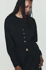 Chaqueta de cuello redondo y manga larga con pliegues, de color negro, con falsos bolsillos de vivo en el delantero y cierre frontal con botones.