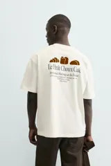 Camiseta de corte relaxed fit, color blanco roto, con cuello redondo y manga corta. Presenta un pequeño estampado en el pecho con un dibujo lineal y texto.