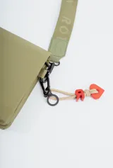 Bandolera de diseño rectangular en color verde oliva, con solapa frontal y logo de la marca grabado. Incluye una correa larga ajustable con el nombre de la marca tejido y un charm decorativo con forma de corazón y letra R en color rojo.