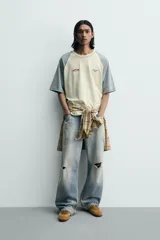 Camiseta de corte relajado con mangas ranglán en color gris y cuerpo en color beige. Presenta cuello redondo, costuras a contraste y detalles de parches y bordados en el pecho.