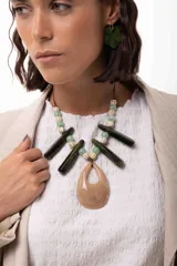 Collar de diseño artesanal con un medallón central en forma de gota color beige marmolado, acompañado de bastones alargados en color verde oscuro y cuentas esféricas en tonos verdes y beige. El diseño se completa con un cierre de tiento regulable.