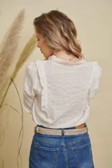 Camisa blanca de manga larga con diseño de volados en los hombros y cuello, confeccionada en tela con textura de rayas verticales y detalles bordados en los bordes. Presenta cierre frontal con botones y puños elásticos con volados.
