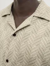 Camisa de manga corta Jack & Jones con cuello resort y cierre de botones. Diseño con estampado de espiga en tonos beige y blanco. Confeccionada en tejido dobby 100% algodón.