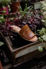 Sandalias tipo chinela de cuero, con diseño de banda ancha que combina cuero liso, cuero texturizado y gamuza en tonos marrones. Presenta plantilla con logo grabado.