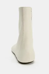 Botín plano de piel en color blanco roto, con diseño minimalista de caña baja, punta redonda y costuras decorativas. Cuenta con cierre mediante cremallera lateral y suela plana.