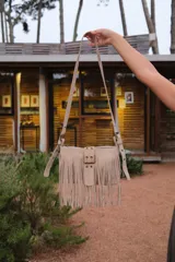 Cartera tipo mini bag de gamuza color marrón, con diseño de flecos largos en todo el frente. Presenta una tira central decorativa con hebilla metálica y herrajes en tono bronce. Incluye correa ajustable y cierre superior.