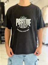 Remera negra con efecto gastado (acid wash), de corte oversize, con estampa frontal en blanco y celeste con la leyenda 'Stay Positive' y diseño gráfico de un globo terráqueo.