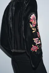 Chaqueta abierta de terciopelo negro con cuello redondo y mangas largas acampanadas. Presenta bordados florales en tonos rosados y verdes, con aplicaciones de abalorios y detalles dorados en el cuello y la parte frontal.