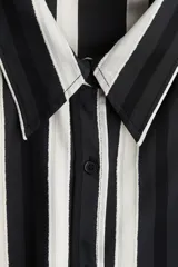 Camisa de manga larga con cuello y botones frontales, hombros caídos y canesú en la espalda. Presenta un patrón de rayas verticales en tonos negro, gris y blanco, con ribete blanco en el cuello.