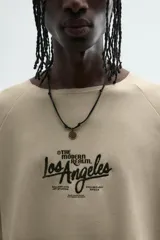 Camiseta de hombre, corte relaxed fit, color beige claro con textura. Presenta cuello redondo amplio y manga corta. Estampado frontal en negro con texto que incluye "Los Angeles".
