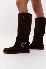 Bota de caña alta confeccionada en gamuza color marrón, con diseño de calce directo y caña flexible que permite drapeado. Presenta dos cinturones desmontables con herrajes metálicos en la zona del tobillo y la parte superior de la caña, con taco bajo y suela de cuero.