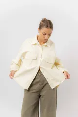 Sobrecamisa de gabardina liviana color crema, con corte oversize, cuello clásico, cierre frontal con botones a presión y dos bolsillos delanteros con solapa.