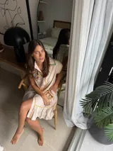 Vestido corto beige de algodón con cuello camisero y cierre frontal de botones. Presenta mangas cortas y detalles bordados a contratono en la zona del pecho y en los bolsillos, además de un volado en el ruedo.