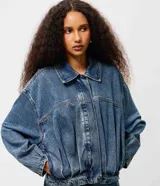 Campera de jean azul con pliegues frontales y posteriores, cuello clásico, cierre frontal embutido y mangas largas. Presenta un diseño de silueta levemente ajustada con terminaciones simples en puños y bajo.