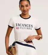 Blusa baby look de algodón color blanco, con cuello redondo y mangas cortas con ribetes azul marino. Presenta un estampado frontal con el texto "VACANCES SANTORINI" en azul marino y detalles florales en rosado.