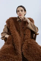 Chaleco largo reversible de diseño versátil. Por un lado, presenta una textura de peluche sintético en color marrón, y por el otro, un acabado liso. Cuenta con cierre frontal de botones y corte recto.