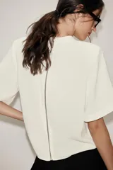 Camisa de cuello subido y manga corta con hombreras. Presenta un diseño minimalista con cierre de cremallera metálica en la espalda.