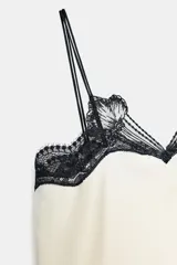 Top de tejido satinado color crema con escote en pico y tirantes finos. Presenta detalles de encaje negro en contraste en el escote y bajo recto.