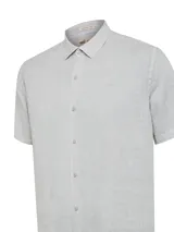 Camisa de manga corta color gris, confeccionada en lino orgánico con cierre frontal de botones, cuello camisero y sutil bordado del logo de la marca en el pecho.