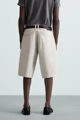 Bermuda color beige claro, de corte ancho y largo, confeccionada en algodón. Presenta cierre frontal con cremallera y botón, bolsillos laterales, bolsillo cerillero frontal y bolsillos traseros de vivo.