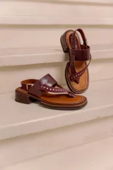 Sandalia de cuero bordó con diseño tipo thong, tiras anchas cruzadas con tachas metálicas doradas, tira con hebilla ajustable en el talón y taco bajo cuadrado con acabado artesanal.
