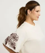 Blusa de corte boxy en color crudo, con cuello redondo y mangas cortas estilo godê. Presenta detalles de bordado floral en tonos marrones en ambas mangas.