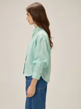 Camisa de lino color verde agua, con cuello clásico, cierre frontal de botones y mangas largas con puños. Presenta detalles de costuras en zigzag en la zona de los hombros y canesú.