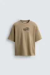 Sudadera de manga corta color beige, con cuello redondo y corte relajado. Presenta un bordado y estampado en el pecho con texto y un diseño floral.