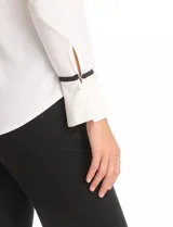 Blusa de crepe negro con ribetes en color beige que recorren el escote, la línea central y las costuras de los hombros. Presenta cuello redondo con abertura en V, mangas largas con puños abiertos y detalle de botón metálico, y ruedo redondeado.