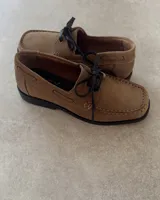 Zapato náutico de cuero vacuno color tostado, con cordones negros y suela plana de goma negra.