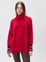 Pullover rojo de cuello alto, corte holgado, mangas largas y puños acanalados en tono burdeos con rayas blancas. Presenta un pequeño logo bordado en el pecho.