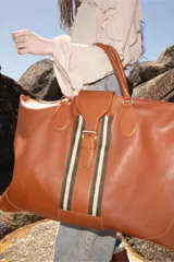Bolso de viaje tamaño XL confeccionado en cuero color suela. Presenta una franja central de cinta tejida en tonos verde y beige con cierre de hebilla metálica. Cuenta con doble correa de hombro y cierre principal.