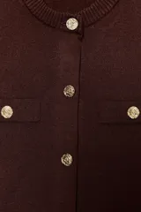 Chaqueta de punto color marrón oscuro, con cuello redondo, manga corta y cierre frontal de botones dorados. Presenta dos bolsillos falsos con botón dorado en la parte delantera superior.