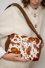 Cartera de mano tipo sobre confeccionada en cuero vacuno con pelo, presentando un diseño estampado con manchas blancas sobre fondo marrón. Posee detalles en gamuza marrón en los laterales y una placa metálica con el logo de la marca en el frente.
