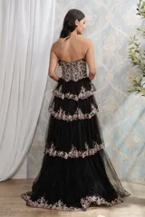 Vestido largo de fiesta color negro, con escote corazón y corset estructurado. Presenta falda de tul con volados escalonados, tajo lateral y bordados florales brillantes en tono dorado.