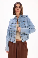 Chaqueta de denim de inspiración napoleónica, confeccionada en jean rígido celeste con lavado medio. Presenta cuello mao, hombreras con presillas y múltiples filas de tiras horizontales decoradas con botones metálicos grabados. De corte recto y mangas largas con puños abotonados.
