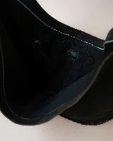 Bolso tipo hobo de gran tamaño, confeccionado en gamuza color marrón oscuro. Presenta un diseño artesanal con costuras visibles y detalles de tientos en el cuerpo, asa integrada con pespuntes a mano y cierre superior.